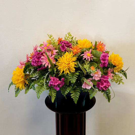 Posies Flowers & Fashion Inc.'s 3 - Tombstone Saddle (60 cm) showcases bright yellow, pink, and purple flowers with green fern leaves in a black vase—a lovely tribute inspired by silk cemetery arrangements.