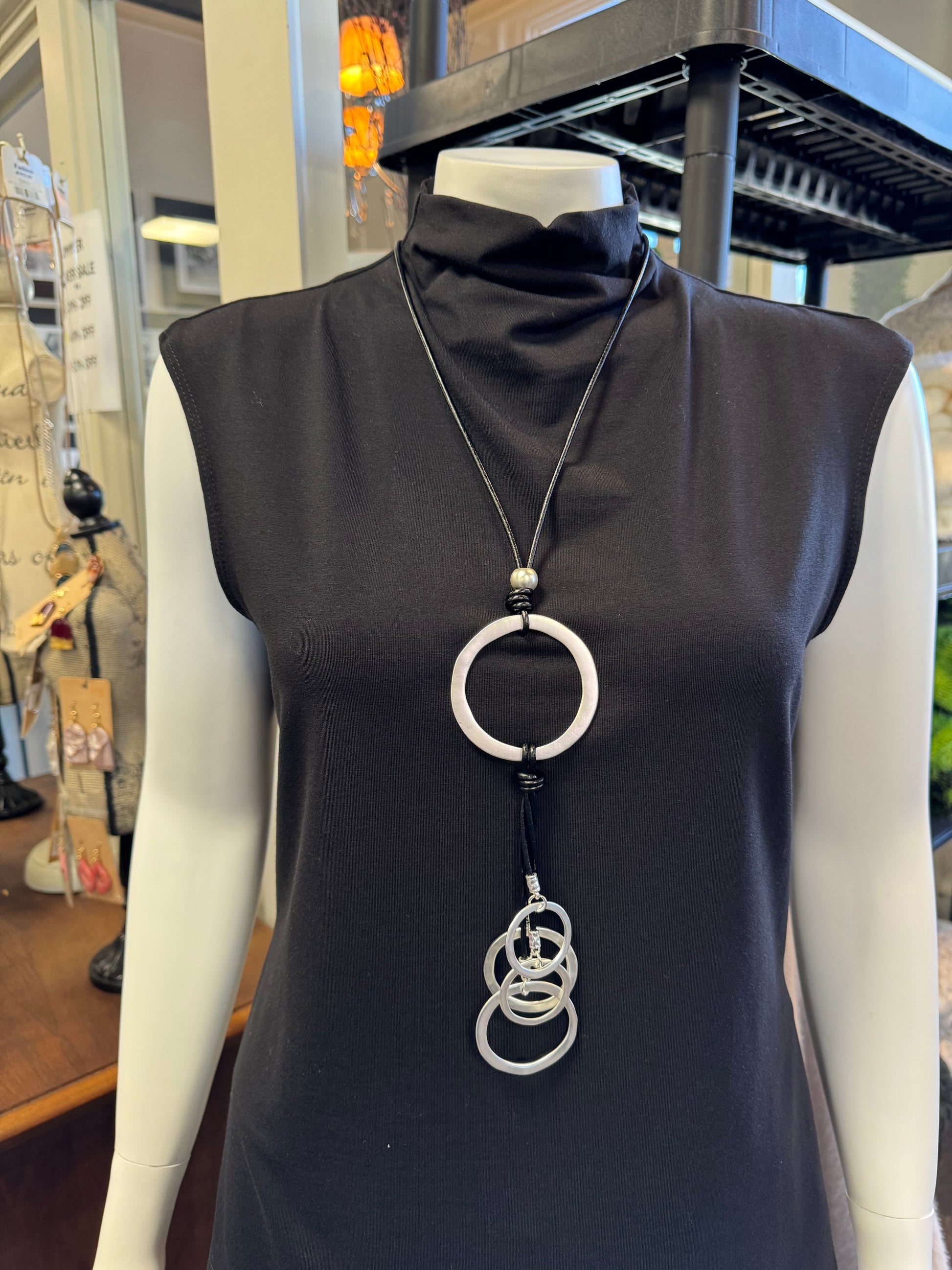 A mannequin showcases a sleeveless black turtleneck dress paired with a striking fashion accessory: the Sofia 3 Necklace by Elise Accessories, featuring interlocking silver rings. The background features a decorative stand and wooden tabletop elements, enhancing this elegant display of jewelry.