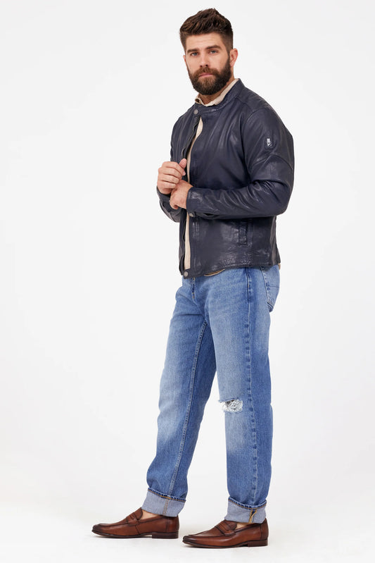 A bearded man wearing a MAURITIUS LEATHER Solvic CF Leather Jacket in navy over a white shirt, paired with light blue rolled-up jeans and brown loafers. He stands against a plain white background, holding the jacket's lapels with both hands while looking confidently towards the camera in his best-selling ensemble.