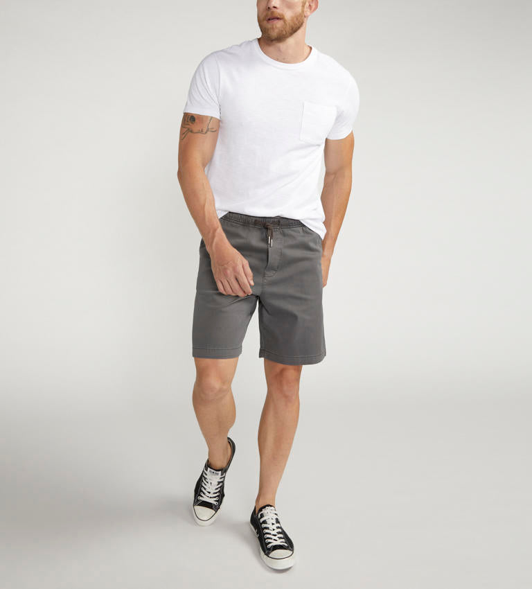 A man is standing while wearing a white short-sleeve t-shirt, Silver Jeans Co.'s PULL-ON CHINO SHORT DARK GREY Style M52155CBT639, and black sneakers with white laces. He has a tattoo on his right arm and a beard. His left hand is in his pocket, and he is looking to his left. The background is plain and light-colored.