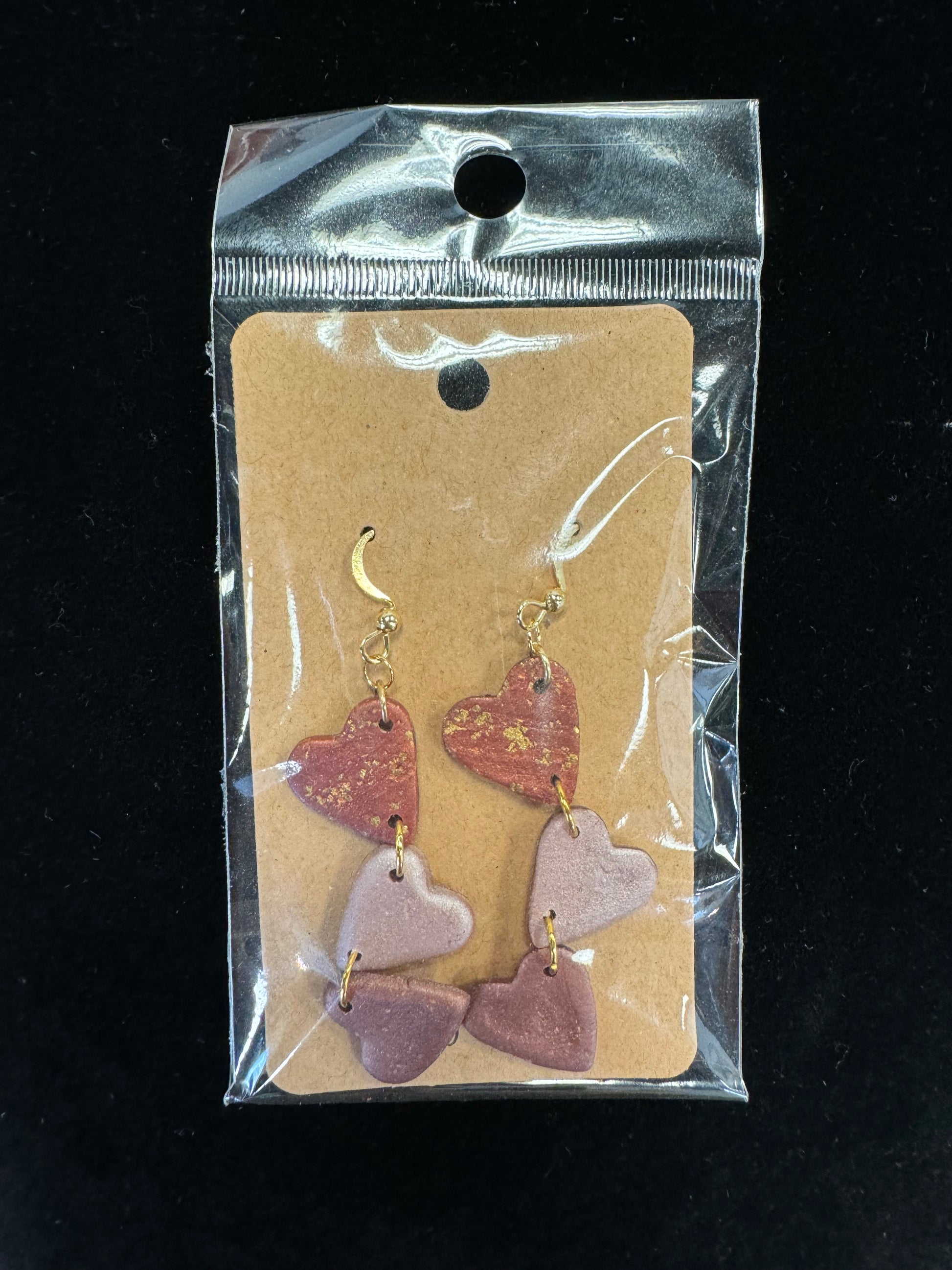 Introducing the Ro&Jo Earrings 425 by RO&JO: a pair of unique high-quality earrings displayed on a beige card inside a clear plastic bag. Each handmade earring features three heart-shaped charms in shades of red and pink, connected in a vertical line, complemented with gold hooks for fastening.