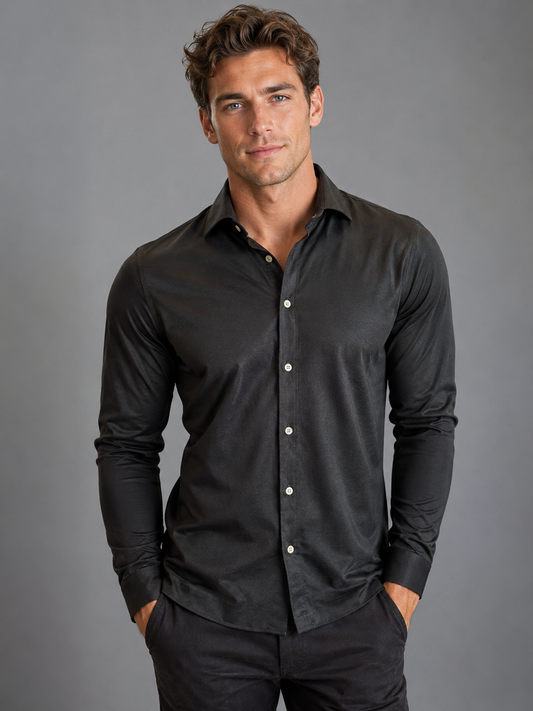 A man with wavy brown hair wears a Paul Miranda Cotton Shirt Black and dark pants, standing against a plain gray background with his hands in his pockets, looking at the camera.