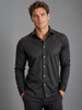 A man with wavy brown hair wears a Paul Miranda Cotton Shirt Black and dark pants, standing against a plain gray background with his hands in his pockets, looking at the camera.