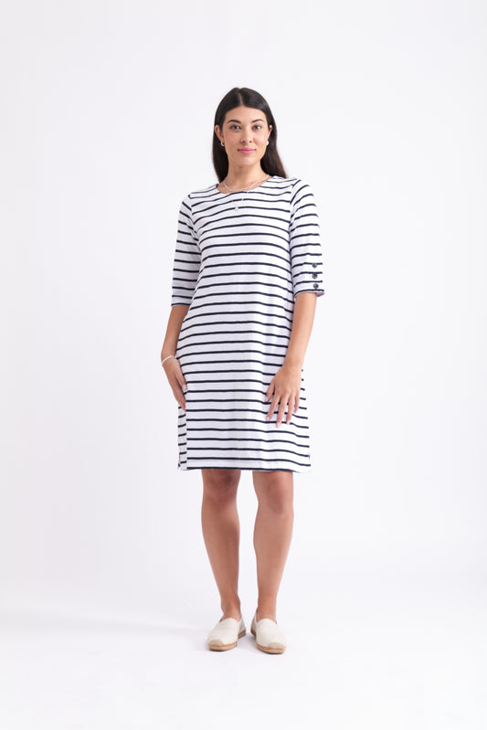 A woman stands against a white background wearing the FOIL Oh Buoy Dress FO7952, a 100% cotton, knee-length summer dress with navy blue horizontal stripes and elbow-length sleeves. She pairs it with beige slip-on shoes and has straight, dark hair.