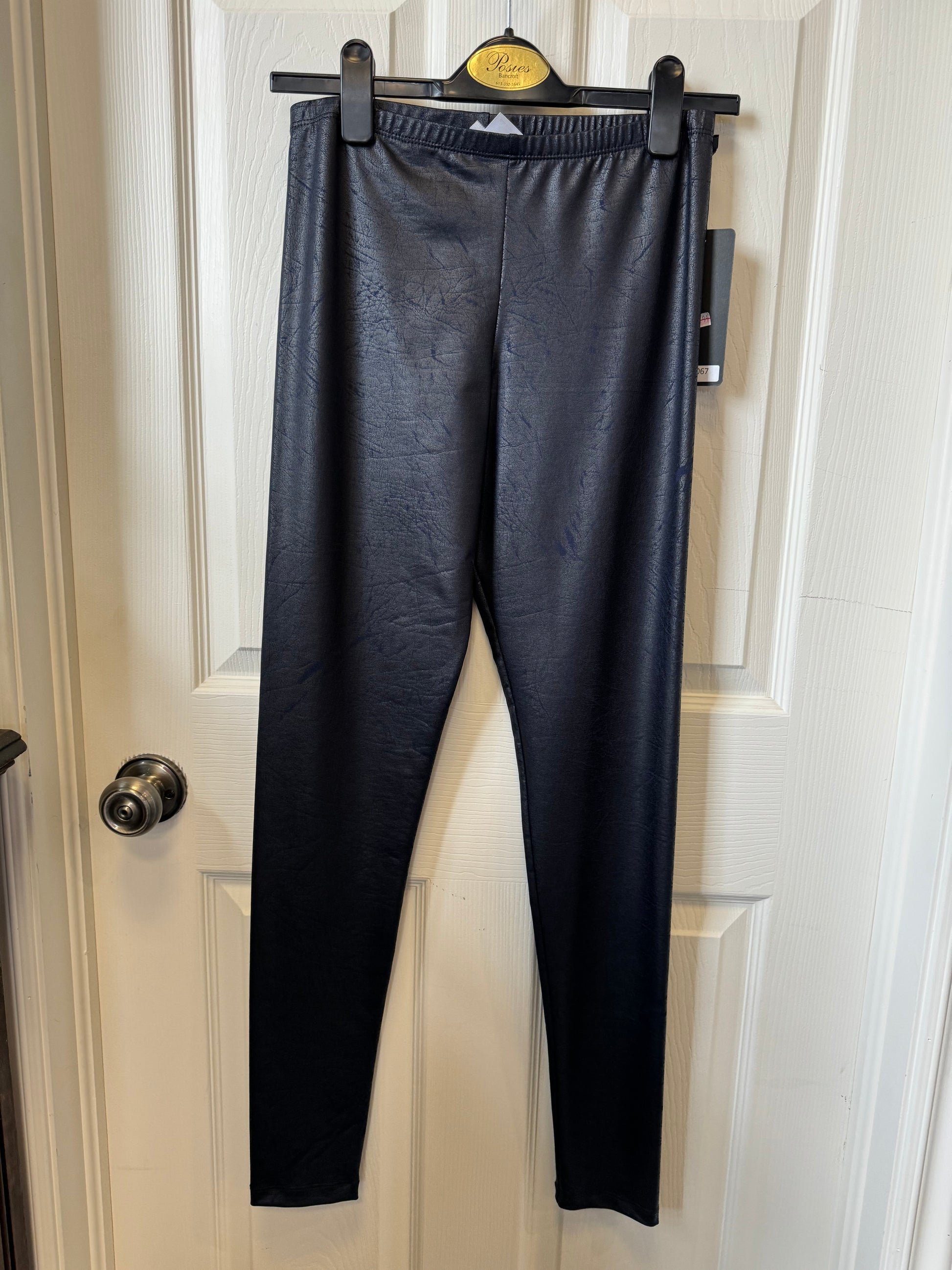 A pair of Dark Navy Leggings by Tribal hang on a black hanger against a white door. The tag on the leggings reads "Artex Fashions - Dark Navy Legging 112-9067" and provides product details. The door has a silver-colored doorknob, and the overall indoor setting promises ultimate comfort and style.