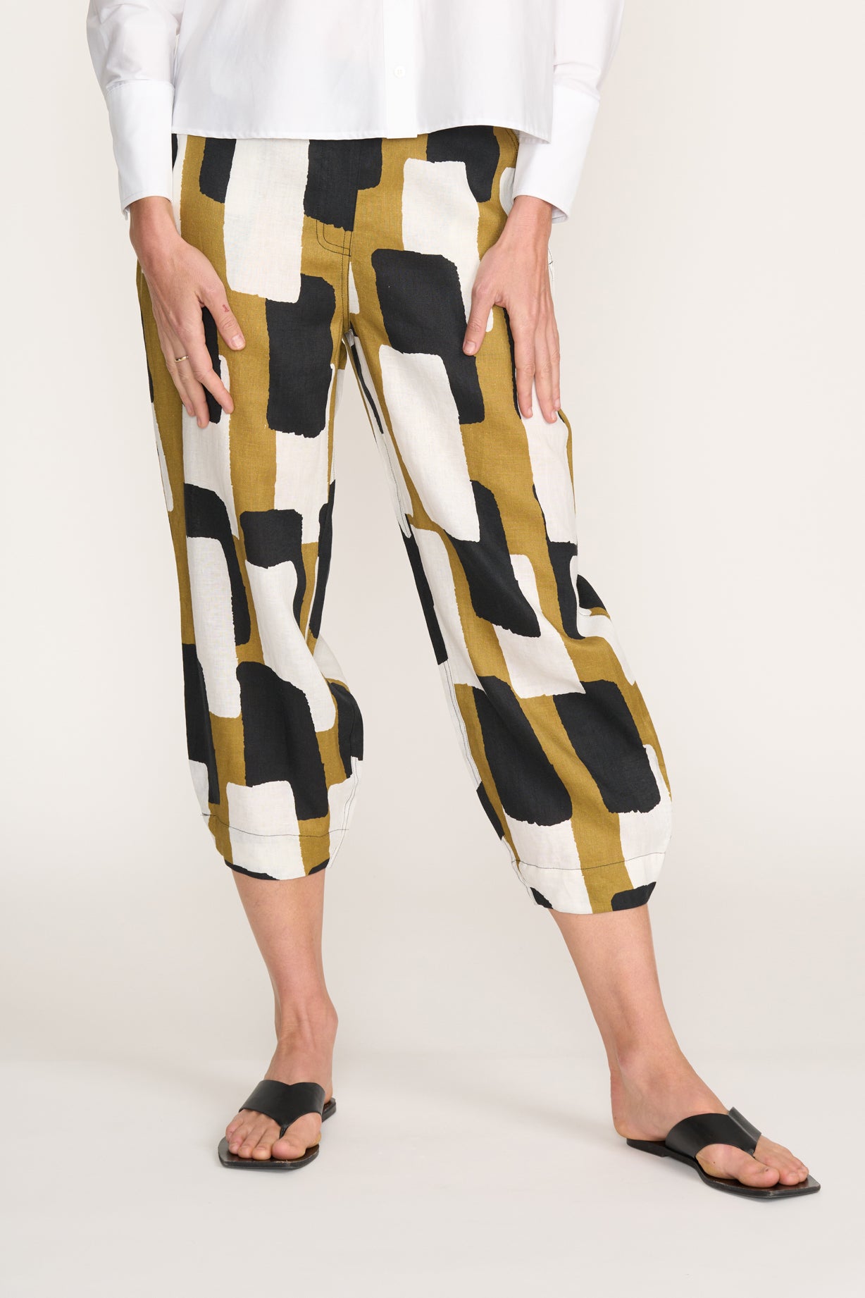 Someone wears the FOIL Jubilant Pant Art Box FO8363—lightweight linen cropped pants in a bold mustard yellow, black, and white print—paired with a white shirt and black sandals against a plain light background.