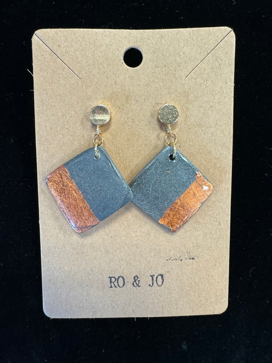 A distinctive pair of handmade Ro&Jo Earrings 427 is presented on a brown card labeled "RO&JO." Each earring has a round gold-colored stud attached to a diamond-shaped pendant made from polymer clay, showcasing gray and metallic copper sections. The background is black.