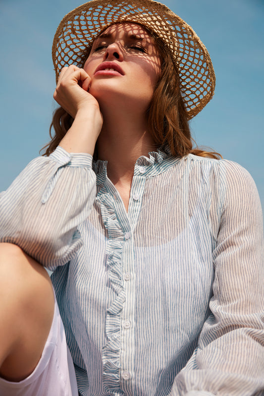 Wearing a straw hat and the effortlessly stylish Top 6324 - SUNDAY by SUNDAY, a person with a thoughtful gaze sits outdoors under the clear blue sky, one hand resting gently on their cheek.