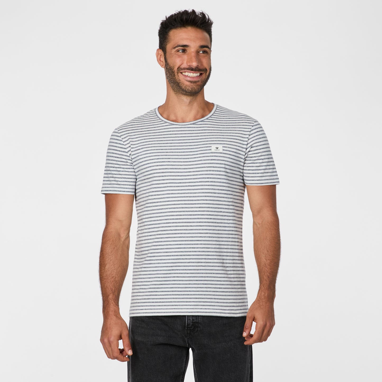A man with short dark hair and a beard smiles in the RAGWEAR Paollo Stripe Gots 2612-15040 Navy men’s t-shirt, paired with black jeans, standing against a plain white background.