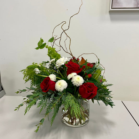 The Clean Winter Vase Arrangement by Posies Flowers & Fashion embodies the spirit of a winter wonderland with its stunning combination of red roses, white chrysanthemums, and lush green foliage presented in a sleek clear glass vase. Curly branches elevate the arrangement with added height and texture, making it an elegant addition to your home decor. It looks particularly striking against a plain white wall on a light-colored table.