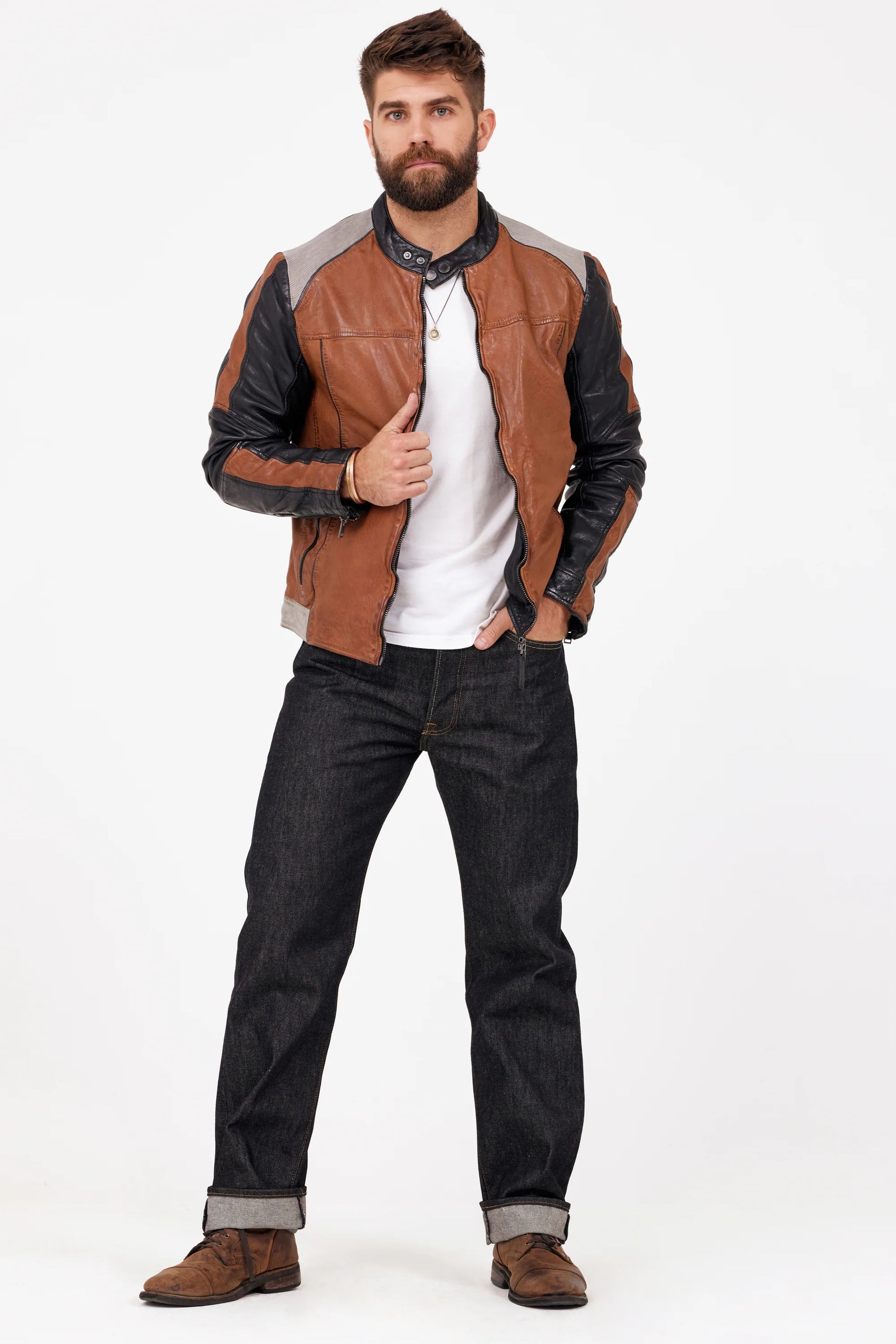 A man with a beard stands confidently against a white background. He is wearing the Myron RF Leather Jacket in Cognac from MAURITIUS LEATHER, paired with a white shirt, dark blue jeans, and brown suede shoes. His hands are in his pockets, and he looks directly at the camera.