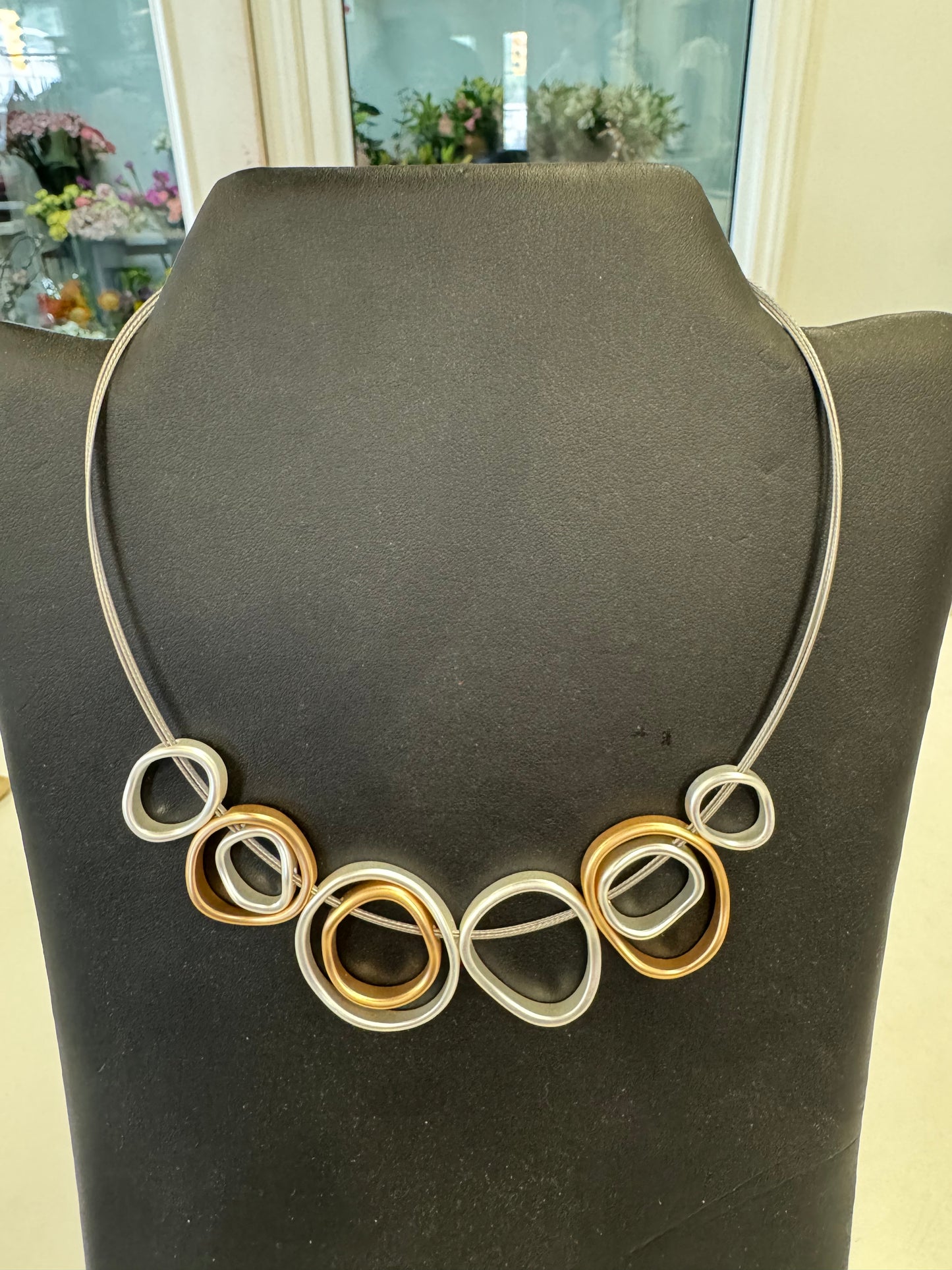 A Loops necklace from Posies Flowers & Fashion Inc., displayed on a black mannequin, features a series of interlocking, abstract circular and oval shapes in silver and gold tones, creating a modern, artistic design. The background reveals a blurred room with various objects, possibly a store. This piece exemplifies elegant jewelry at its finest.