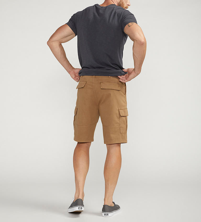 A man stands against a plain background wearing a dark gray t-shirt with a chest pocket, relaxed fit CARGO SHORT DESERT styled by Silver Jeans Co. (product code: M53151CBT639), and black sneakers with white soles. He appears to be adjusting his shirt with his right hand.