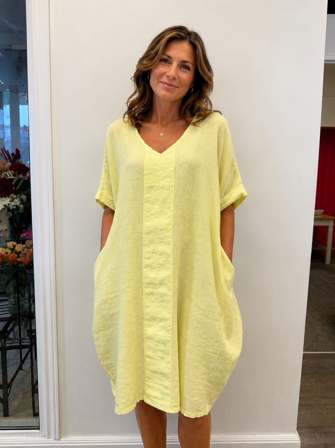 A woman with wavy brown hair smiles indoors beside flowers, wearing the Linen Dress Yellow 6-SS26219-9 by ETERNELLE—a loose-fit summer linen dress with pockets, made in Italy, perfectly styled against a plain white wall.