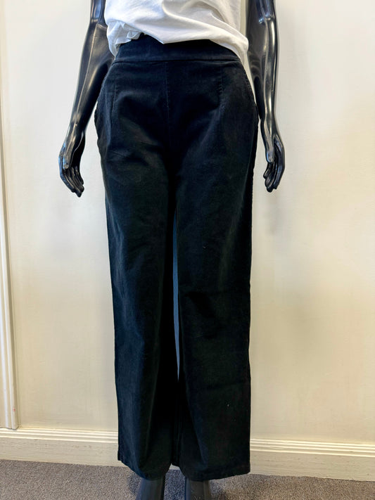 A mannequin with black, shiny arms showcases the EKRU - BRUSHED CORD PANT - Nero, featuring convenient pockets, standing against a plain white wall. The mannequin also wears a white shirt from EKRU europe, its bottom part clearly visible. The beige carpeted floor is seen at the base.