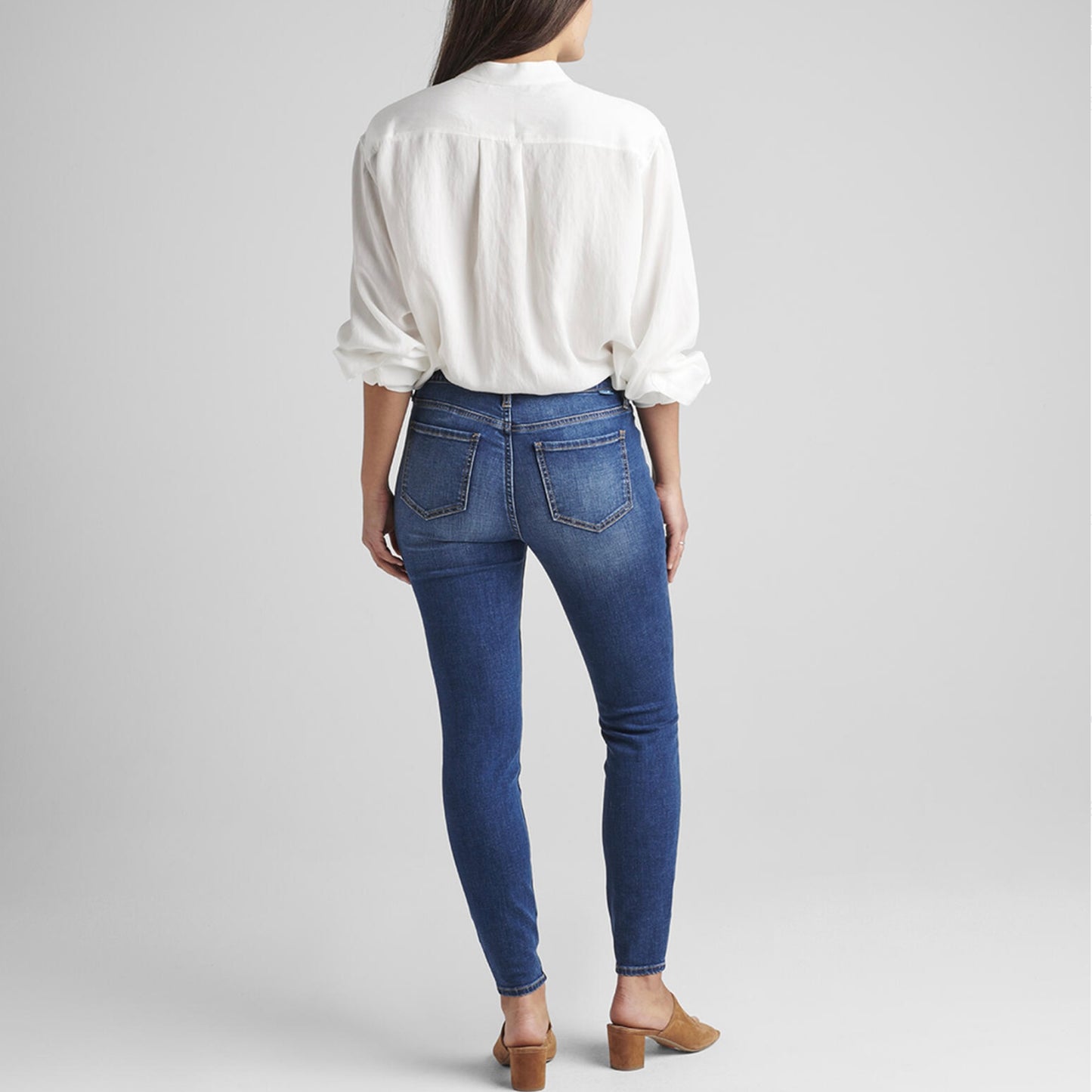 A person stands against a plain background, wearing a white, long-sleeved shirt with front pockets, tucked into Cecilia Mid Rise Skinny Jeans featuring Best Kept Secret technology by JAG. They are also wearing brown, open-toe heeled sandals. One hand is tucked into the jeans pocket while the other hangs at their side.