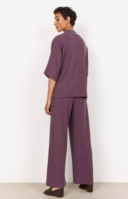 Standing against a plain white background, a person with short curly hair showcases the SC-ISEL 1 Blouse from SoyaConcept. The two-piece purple outfit, made from LENZING™ ECOVERO™, features vertical stripes. Facing away from the camera, they highlight the back of the loose-fitting top and wide-legged pants, creating a minimalist look.