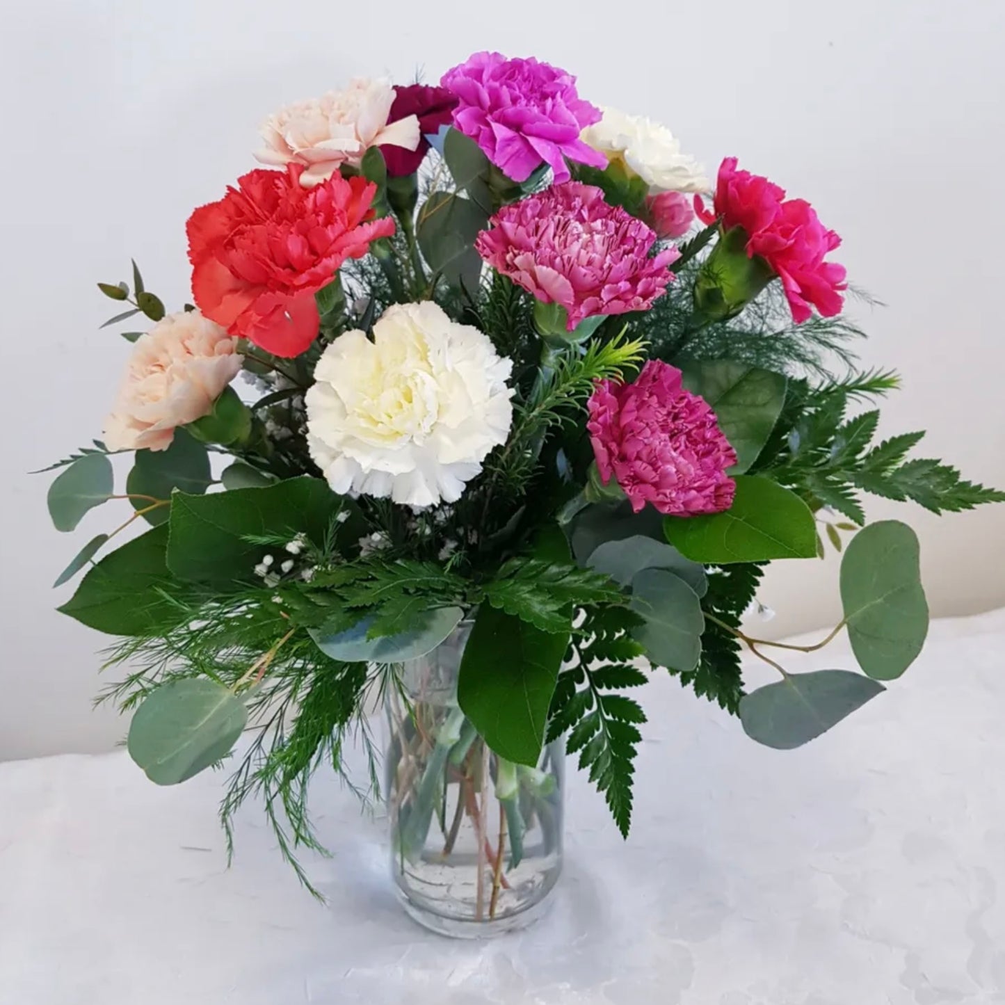 The "Carnation Sensation" bouquet from Posies Flowers & Fashion features a clear glass vase filled with a lively assortment of red, white, pink, and purple carnations. Green foliage enhances the flowers, adding depth and texture to this vibrant arrangement set against a plain white background.