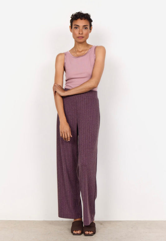 A person with short curly hair is standing against a plain white background. They are wearing a sleeveless pink top and the SC-ISEL 1 Pants by SoyaConcept, which are high-waisted, wide-leg purple pants featuring a subtle pinstripe design. Made from recycled polyester, the pants complement their arms slightly crossed as they look at the camera.