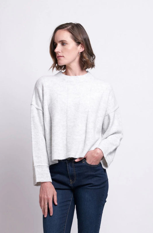 A person with short, wavy brown hair stands against a plain white background. They are wearing a stylish, loose-fitting light grey FOIL Tread Softly Sweater and dark blue jeans. Their left hand is in their pocket, and they look to their left with a neutral expression.