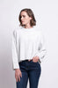 A person with short, wavy brown hair stands against a plain white background. They are wearing a stylish, loose-fitting light grey FOIL Tread Softly Sweater and dark blue jeans. Their left hand is in their pocket, and they look to their left with a neutral expression.