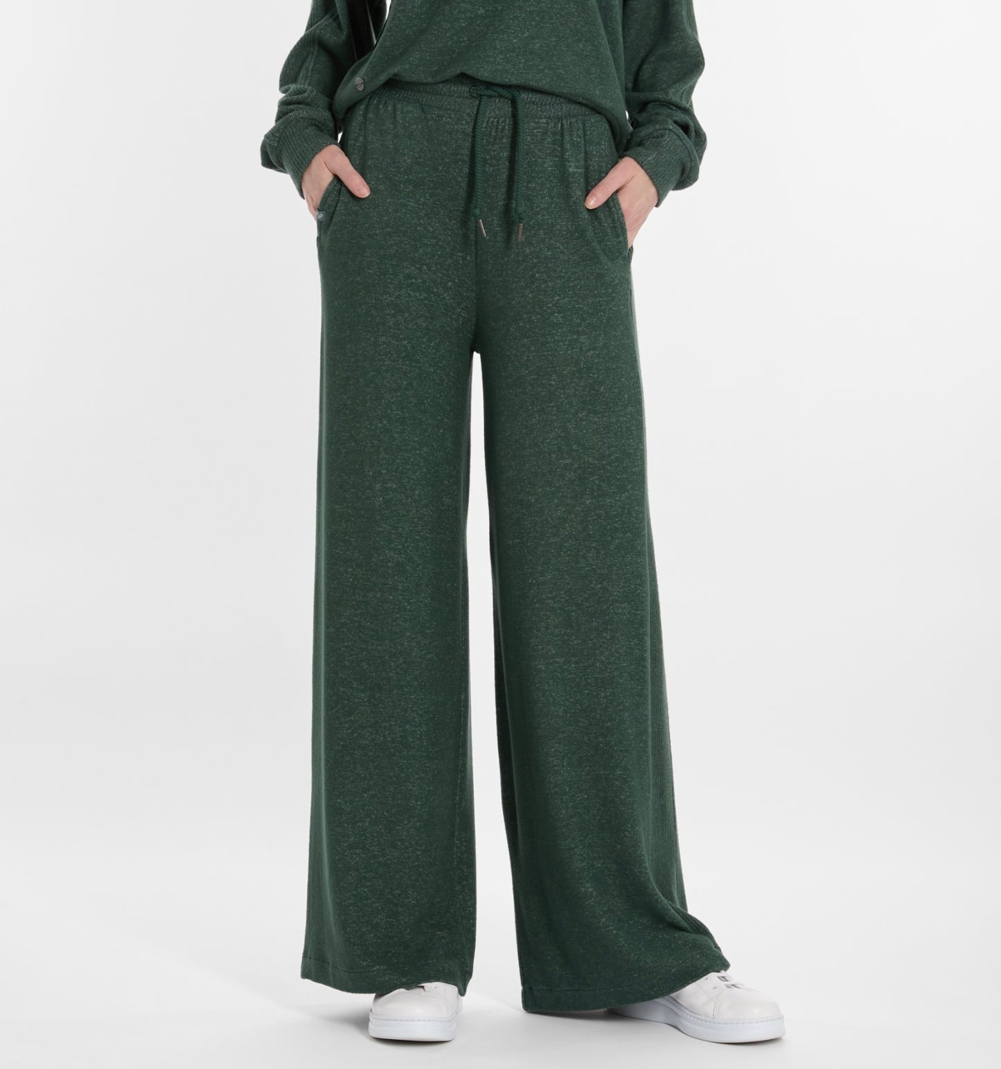 A person, shown from the waist down, wears RAGWEAR JEGGY Women’s Pants in pine green with a matching top and classic white sneakers; their hands rest casually in their pockets.