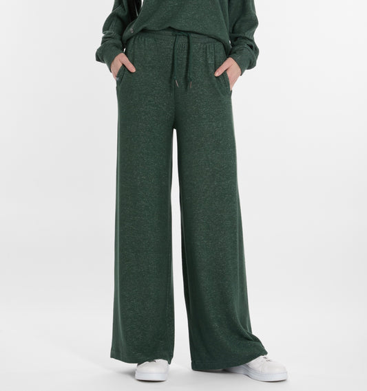 A person, shown from the waist down, wears RAGWEAR JEGGY Women’s Pants in pine green with a matching top and classic white sneakers; their hands rest casually in their pockets.