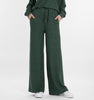 A person, shown from the waist down, wears RAGWEAR JEGGY Women’s Pants in pine green with a matching top and classic white sneakers; their hands rest casually in their pockets.