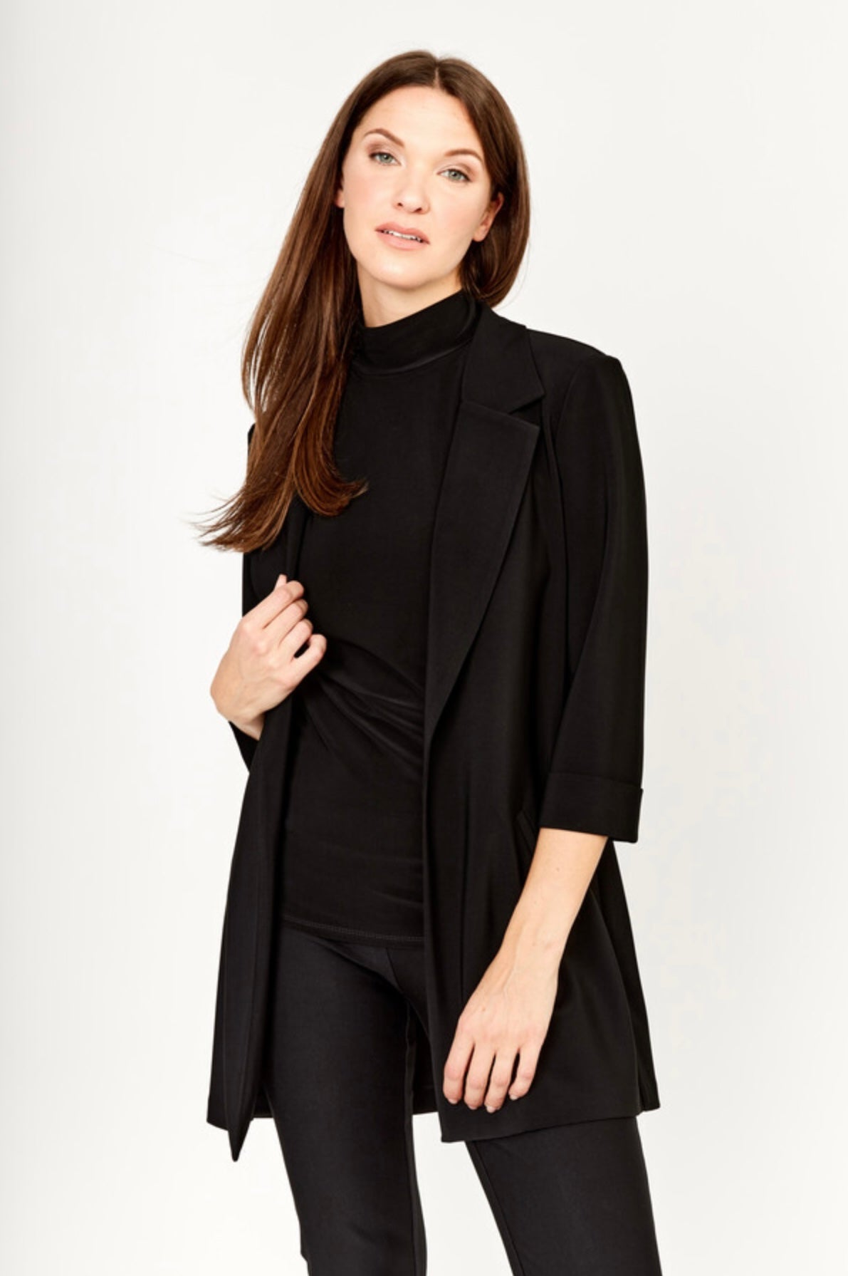 A woman with long brown hair models the Frank Lyman Long Open-Front Jacket in black (Style 236005) as a chic layering piece over a black turtleneck top and black pants, standing against a plain light background.