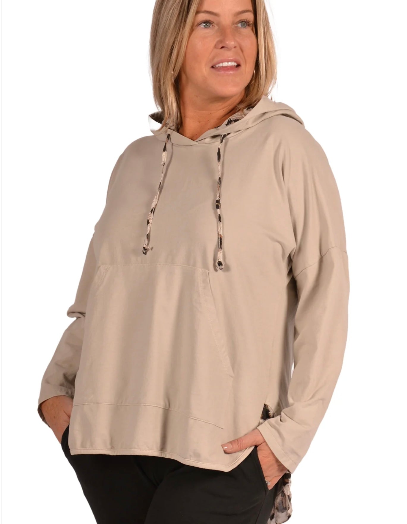 A woman with shoulder-length blonde hair is wearing a beige ITM5008BE-Hoodie Catherine Lilywhite from Catherine Lillywhite and black pants. She is standing with her left hand in the pocket of her sweatshirt, looking off to the side. The background is plain white.