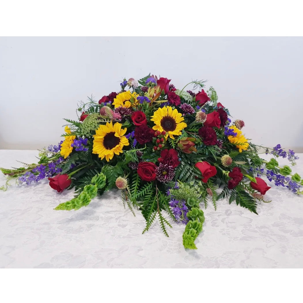 The "Sunflower Dreams - Casket Spray" by Posies Flowers & Fashion features a large, lush white container filled with a cascading arrangement of vibrant yellow sunflowers, striking red roses, and an assortment of purple and green flowers interwoven with various greenery. This elegant display creates a colorful and captivating visual. Availability may vary.