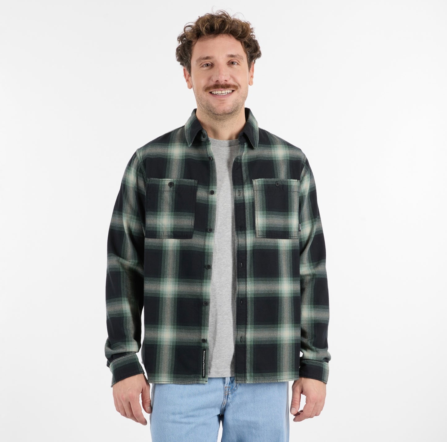 A man with curly brown hair models the RAGWEAR COLLKI Mens Flannel Shirt in green and black over a gray tee and light blue jeans, standing and smiling against a plain white background.