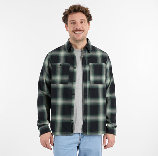A man with curly brown hair models the RAGWEAR COLLKI Mens Flannel Shirt in green and black over a gray tee and light blue jeans, standing and smiling against a plain white background.