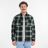 A man with curly brown hair models the RAGWEAR COLLKI Mens Flannel Shirt in green and black over a gray tee and light blue jeans, standing and smiling against a plain white background.