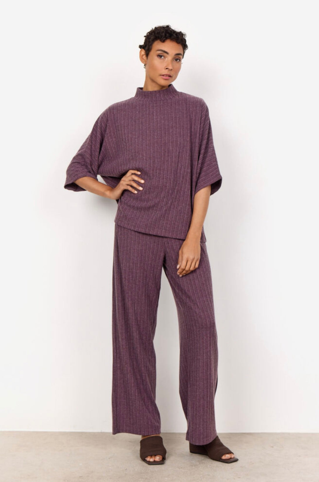 Standing against a plain white background, a person with short curly hair showcases the SC-ISEL 1 Blouse from SoyaConcept. The two-piece purple outfit, made from LENZING™ ECOVERO™, features vertical stripes. Facing away from the camera, they highlight the back of the loose-fitting top and wide-legged pants, creating a minimalist look.