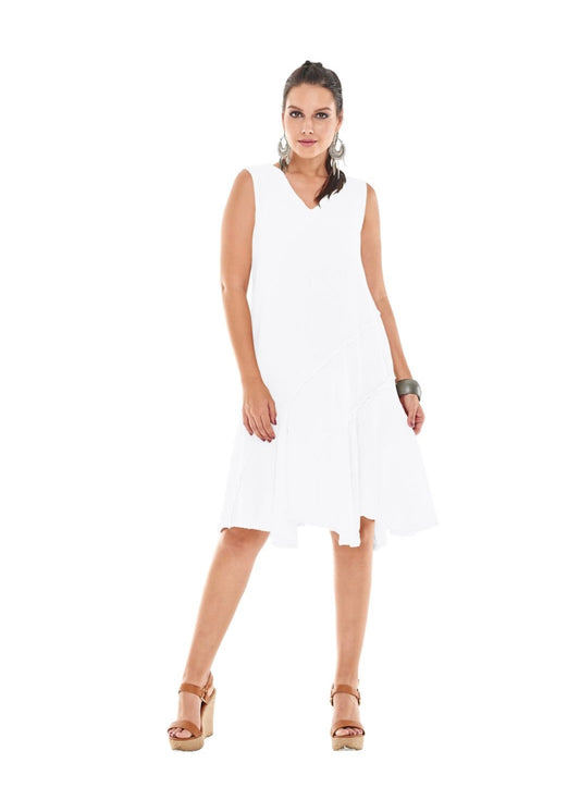 A woman is standing and wearing the TABASCO DRESS SNOW by Oh My Gauze, a sleeveless, white, knee-length gauze dress with a v-neckline. She has a confident pose with one leg slightly bent and is accessorized with hoop earrings, a bracelet, and wedge sandals. The background is plain white.