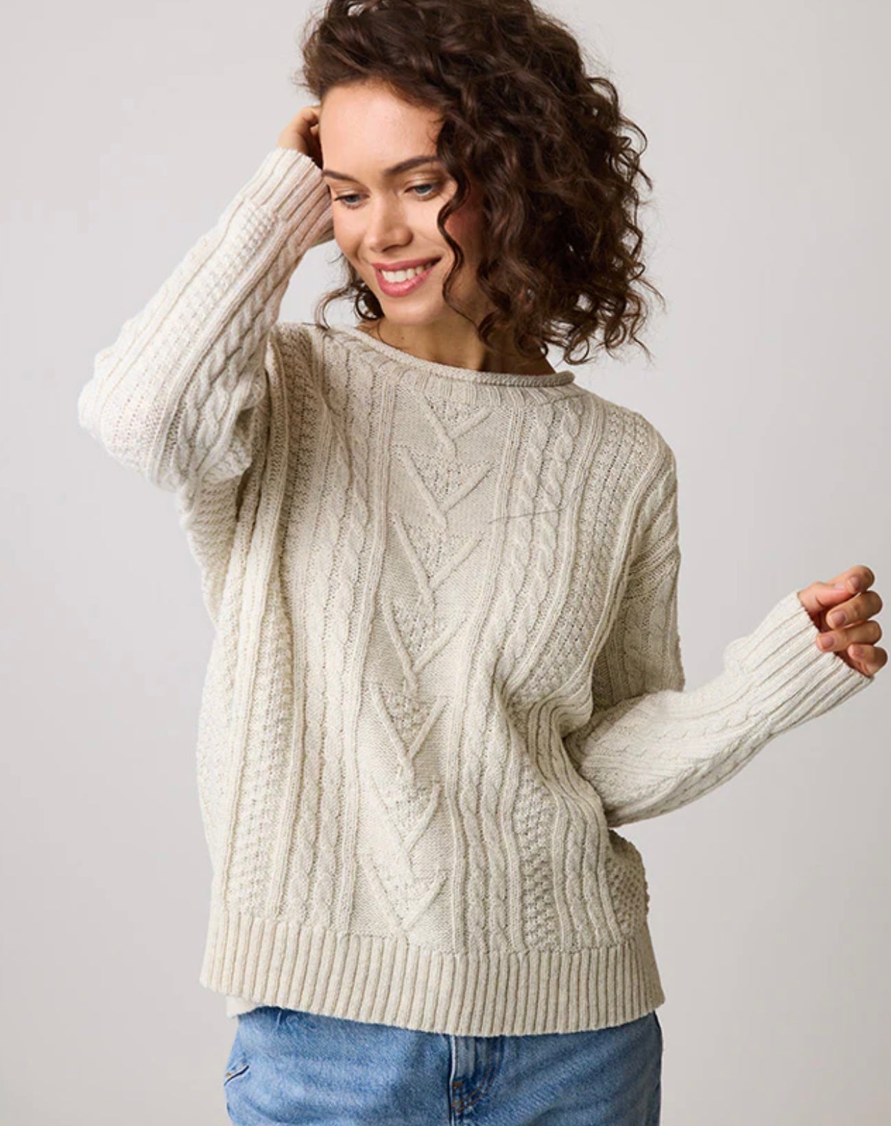 A person with curly hair smiles while wearing the JENNIFER CREW 85142, a cream-colored cable-knit sweater made from eco cotton by Parkhurst, paired with blue jeans. The background is plain and neutral.