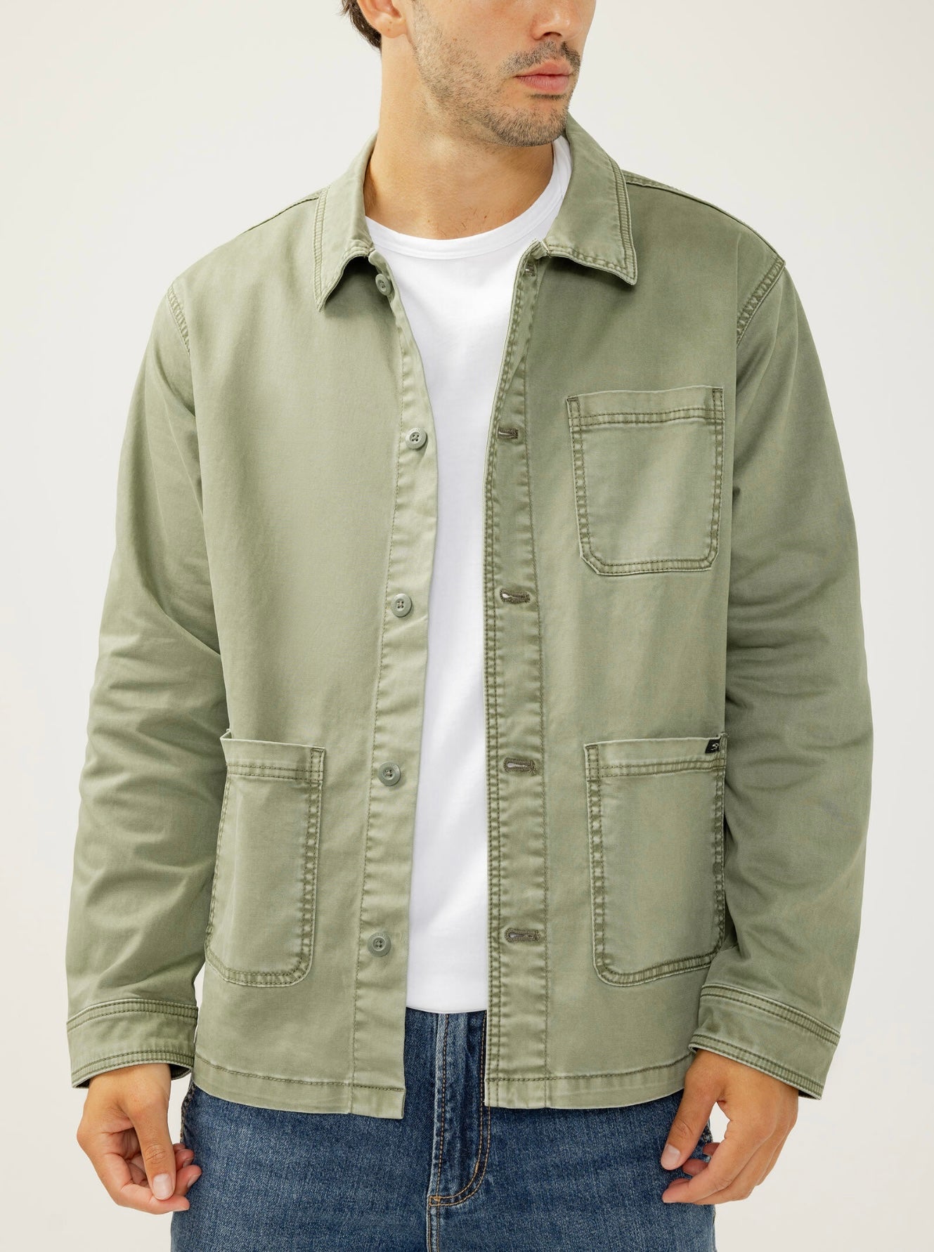 A man wearing the Essential Twill Chore Jacket MJ0028CBT675 by Silver Jeans Co. in light olive green, paired with a white t-shirt and blue jeans, stands against a plain background, his face partially turned away from the camera.