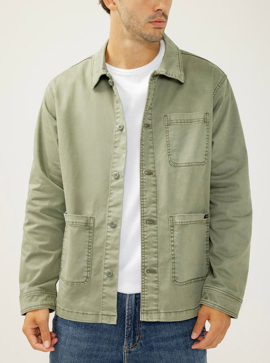 A man wearing the Essential Twill Chore Jacket MJ0028CBT675 by Silver Jeans Co. in light olive green, paired with a white t-shirt and blue jeans, stands against a plain background, his face partially turned away from the camera.