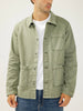A man wearing the Essential Twill Chore Jacket MJ0028CBT675 by Silver Jeans Co. in light olive green, paired with a white t-shirt and blue jeans, stands against a plain background, his face partially turned away from the camera.