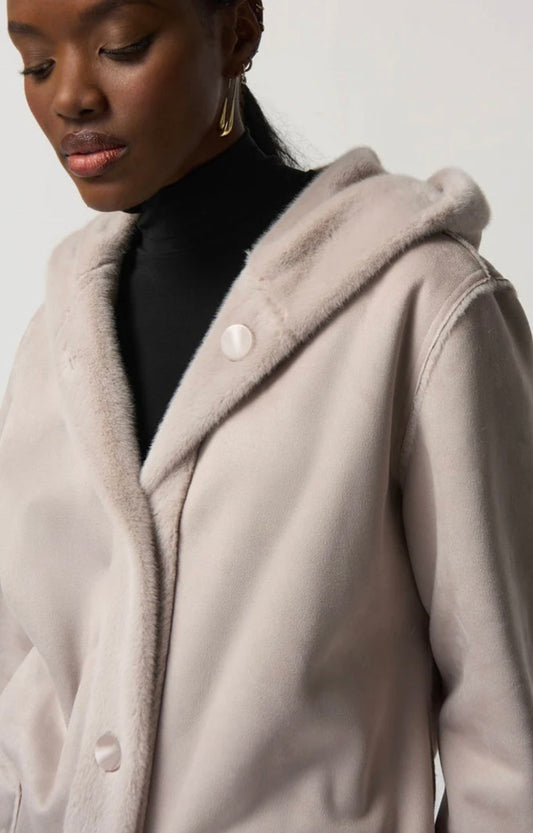 A person wearing a light-colored Joseph Ribkoff Coat 233922, made of faux suede with a fur-lined hood and large buttons. The individual has a thoughtful expression and is dressed in a black turtleneck underneath the coat, epitomizing winter style. The background is plain and light-colored.