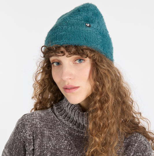 A woman with long curly hair models the teal fuzzy knit YAFFA Hat by RAGWEAR and a textured gray turtleneck, posing against a plain white background.