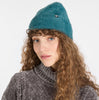 A woman with long curly hair models the teal fuzzy knit YAFFA Hat by RAGWEAR and a textured gray turtleneck, posing against a plain white background.