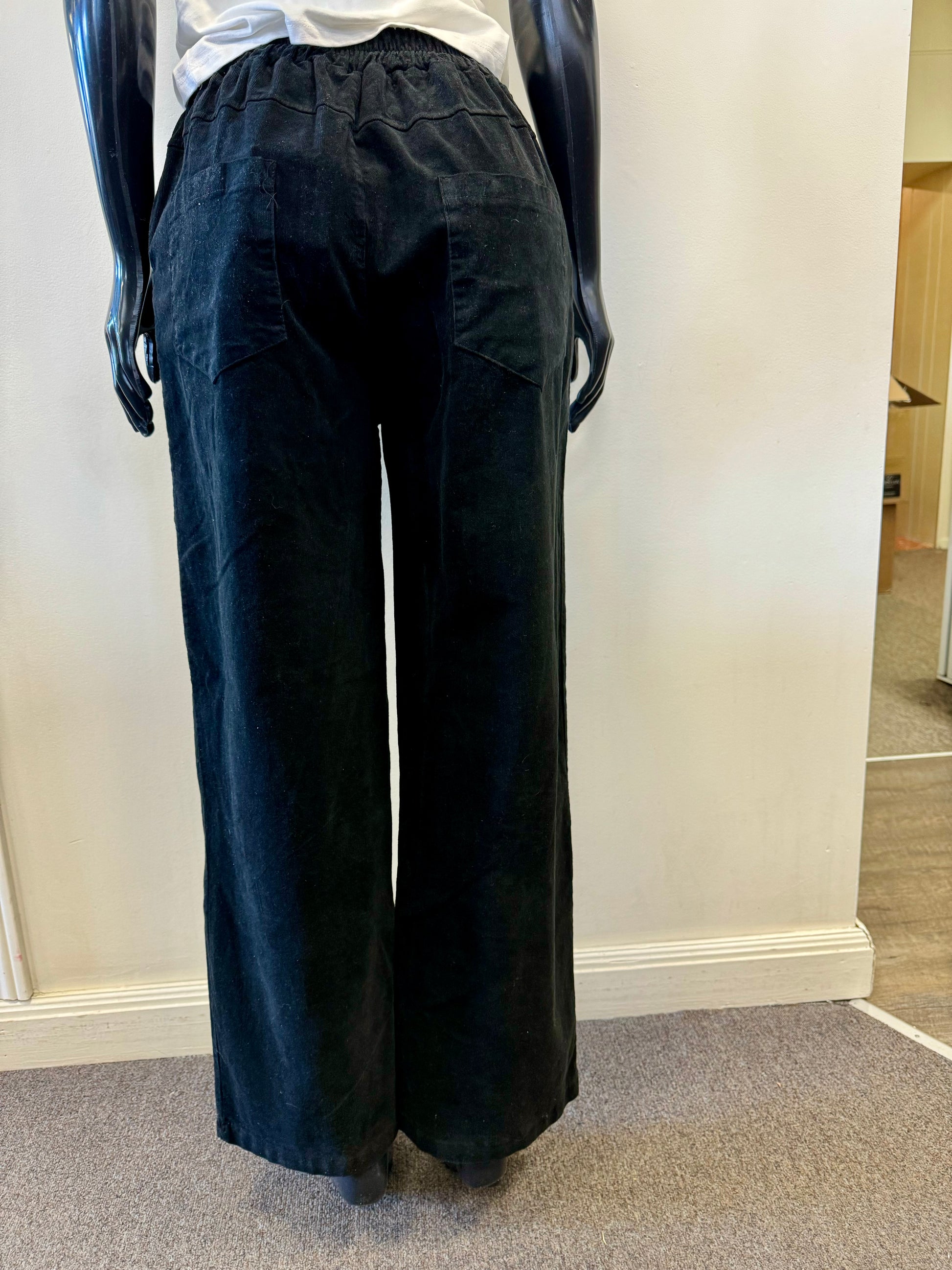 A mannequin with black, shiny arms showcases the EKRU - BRUSHED CORD PANT - Nero, featuring convenient pockets, standing against a plain white wall. The mannequin also wears a white shirt from EKRU europe, its bottom part clearly visible. The beige carpeted floor is seen at the base.