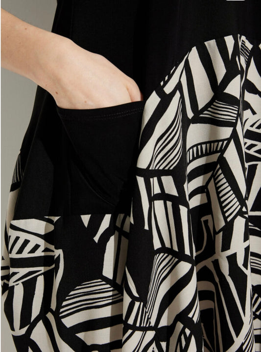 Close-up of a person wearing the Joseph Ribkoff Solid & Printed Dress Style 232112, showcasing its black and white abstract motif. The fabric features geometric designs, and the person's left hand is casually placed inside a side pocket of this work-appropriate dress. The background is neutral and out of focus.