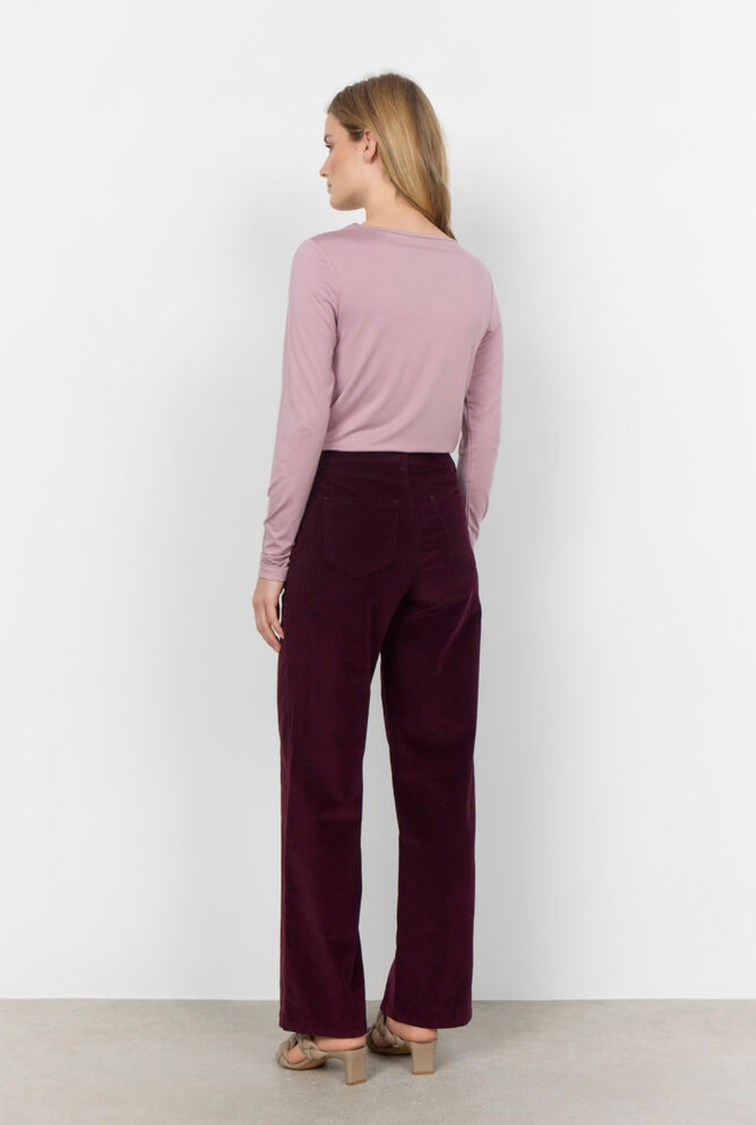 A person wearing a light pink long-sleeve shirt and SoyaConcept's SC-IVALOU 3-C dark purple velvet organic cotton straight-leg pants stands on a light-colored floor. Their feet are partially visible, showing flat, beige braided sandals. The background is plain and light-colored.