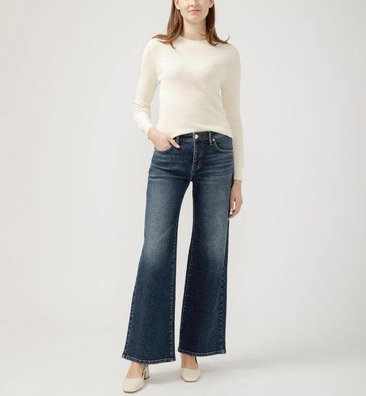 A woman stands against a plain light gray background, wearing a fitted white long-sleeve sweater and the Willow Mid Rise Wide Leg Jeans by JAG, which feature 5-pocket styling. She has one hand in her pocket and is wearing beige heeled shoes.