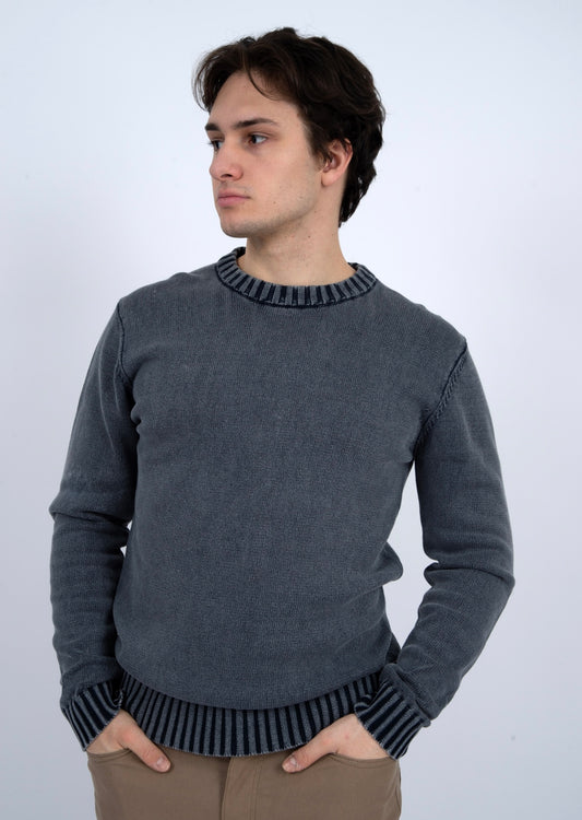 A young man with medium-length brown hair wears the HEDGE CREW SWEATER-NAVY ACID WASH, featuring striped cuffs and collar, standing with hands in pockets and looking to the side against a plain light background.