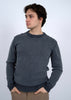 A young man with medium-length brown hair wears the HEDGE CREW SWEATER-NAVY ACID WASH, featuring striped cuffs and collar, standing with hands in pockets and looking to the side against a plain light background.