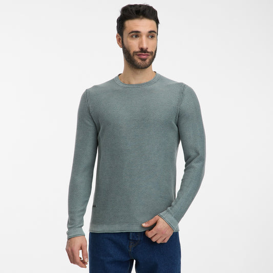 A man with short dark hair and a beard is wearing the TORIEN Mens Flat Knit Shirt by RAGWEAR in light green and blue jeans, standing against a plain white background, looking slightly to the side.
